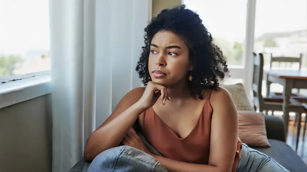 Mulher pensativa olhando pela janela e vivendo excesso de pensamentos no dia a dia